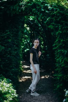 Portrait of one beautiful Caucasian young teenage girl in glasses and sportswear standing on a path with a plant arch in the botanical garden of wonders on a summer sunny day in France, close-up side view with depth of field.