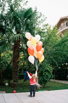 Little girl stands with her hand raised with a bunch of balloons in the garden and looks up. Back view. High quality photo