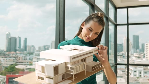 Young smart architect engineer holds architectural model while inspect house model. Professional interior designer checking house construction while standing near window with city view. Tracery