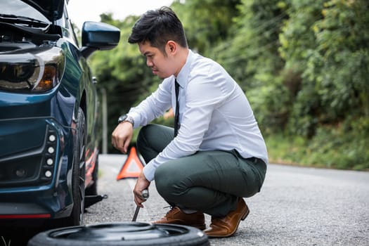Experienced Asian engineer changing car wheel on the highway. Handsome businessman in white shirt watches the repair process. Skilled mechanic providing roadside assistance.