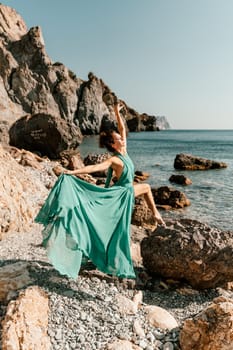 Woman green dress sea. Woman in a long mint dress posing on a beach with rocks on sunny day. Girl on the nature on blue sky background