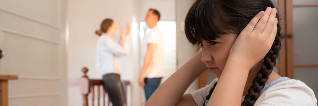 Stressed and unhappy young girl huddle in corner, cover her ears blocking sound of her parent arguing in background. Domestic violence at home and traumatic childhood develop to depression. Synchronos