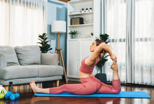 Flexible and dexterity woman in sportswear doing yoga position in meditation posture on exercising mat at home. Healthy gaiety home yoga lifestyle with peaceful mind and serenity.