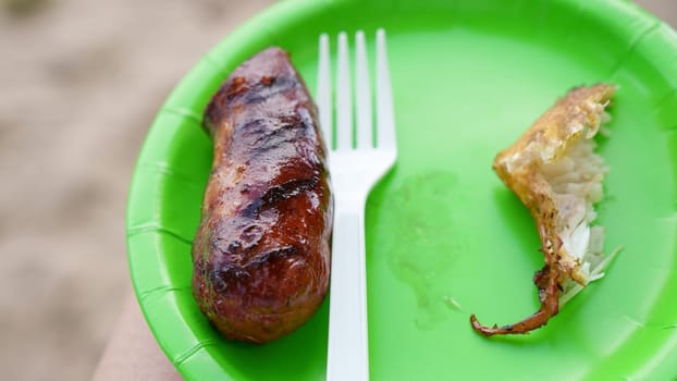 Closeup of a roasted sausage with plastic fork on green paper plate. Camping food