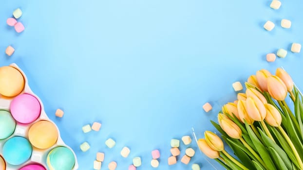 Spring flat lay with fresh yellow tulips colorful eggs on blue background. Easter, Mother day, birthday or Anniversary with copy-space. Sweet rainbow color Easter eggs, small pastel marshmallows