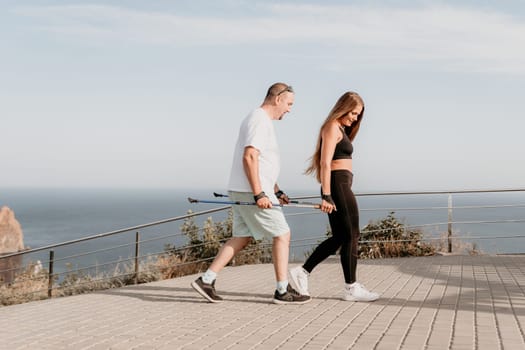 Happy Middle aged couple or friends practicing nordic walking in park near sea. Mature couple with trekking poles walking, practicing Nordic walking outdoors. Aging youthfully and sport concept.