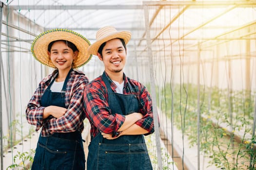 Tomato hydroponic farm delight with Asian couple. Confident farmers crossed arms carrying quality vegetables. Portrait of successful husband and wife in the greenhouse. Farming joy.