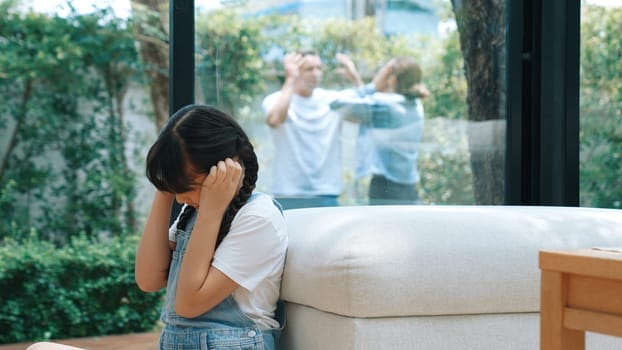 Stressed and unhappy young girl huddle in corner, cover her ears blocking sound of her parent arguing in background. Domestic violence at home and traumatic childhood develop to depression. Synchronos