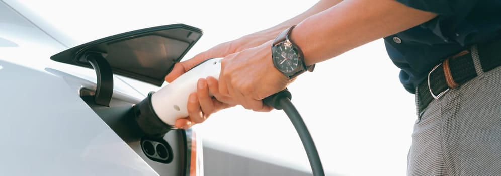 Young man put EV charger to recharge electric car's battery from charging station in city commercial parking lot. Rechargeable EV car for sustainable environmental friendly urban travel. Expedient