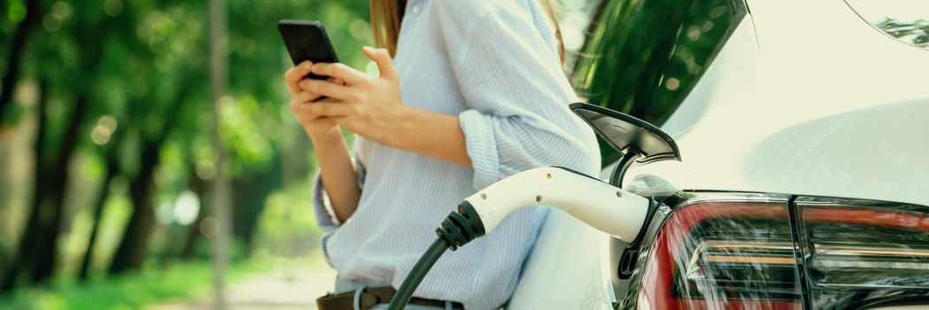 Young woman using smartphone online banking application to pay for electric car battery charging from EV charging station during vacation road trip at national park or summer forest. Panorama Exalt