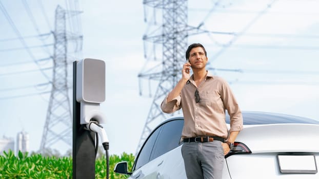 Man talking on the phone while recharge EV car battery at charging station connected to power grid tower electrical as electrical industry for eco friendly car utilization. Panorama Expedient