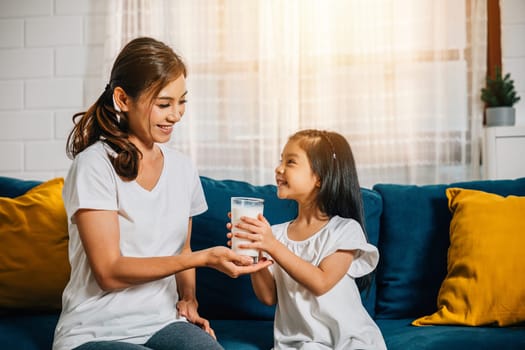 A joyful scene as an Asian mother gives her daughter a glass of milk on the living room sofa. This image radiates affection innocence and the happiness of family breakfasts.