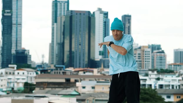 Skilled caucasian B-boy dancer practicing street dancing at rooftop with city sites or urban. Motion shot of young man performing street dance by doing freeze pose. Outdoor sport 2024. Endeavor.