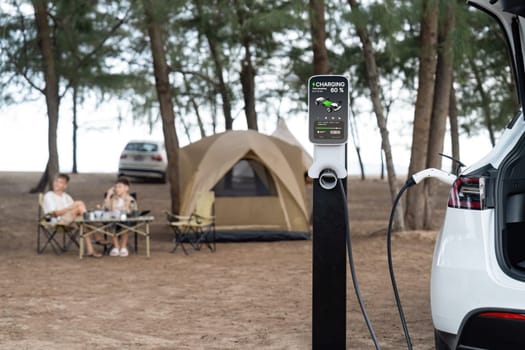 Alternative family vacation trip traveling by the beach with electric car recharging battery from EV charging station with two family kid enjoying the seascape camping background. Perpetual