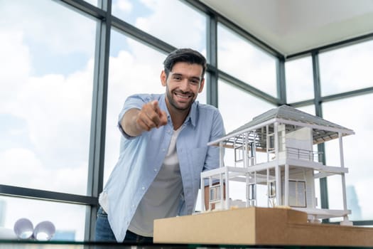 Portrait of smart engineer in casual outfit smiling at camera while holding pencil. Businessman pointing at camera and standing near house model, project plan with sky scraper behind. Tracery.