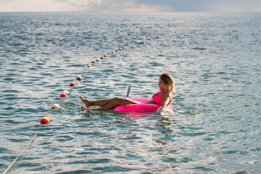 Woman works on laptop in sea. Freelancer, blond woman in sunglases floating on an inflatable big pink donut with a laptop in the sea at sunset. People summer vacation rest lifestyle concept