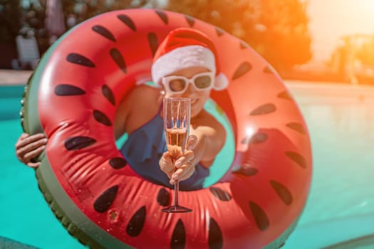 Woman pool Santa hat. A happy woman in a blue bikini, a red and white Santa hat and sunglasses poses near the pool with a glass of champagne standing nearby. Christmas holidays concept