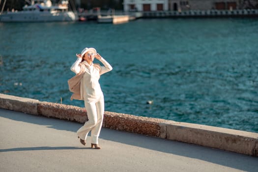 Happy blonde woman in a white suit and hat posing at the camera against the backdrop of the sea.