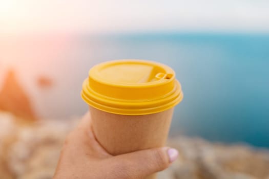 Hand holding Yellow cup with lid, coffee against a backdrop of a blue sky and sea. Illustrating cup and beverage.