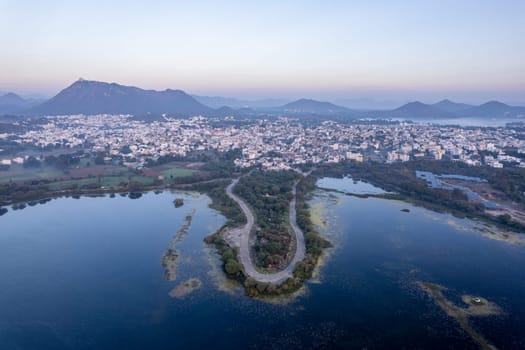 aerial drone shot at dawn dusk with road loop extending into fateh sagar lake with aravalli hills in distance hidden in fog showing cityscape of Udaipur Rajasthan India