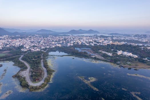 aerial drone shot at dawn dusk with road loop extending into fateh sagar lake with aravalli hills in distance hidden in fog showing cityscape of Udaipur Rajasthan India