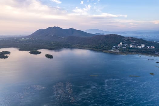 Aerial drone shot dawn dusk sky with aravalli hills with homes, houses at base and blue weed covered waters of Pichola Fateh Sagar lake in Udaipur, Nainital in India