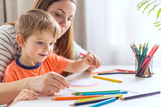 A young boy and his mother share a bonding moment while drawing with colorful pencils at home, with a plant in the background.
