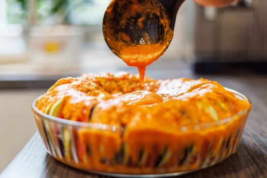 Pouring rich tomato sauce over a beautifully arranged vegetable tian in a glass dish.