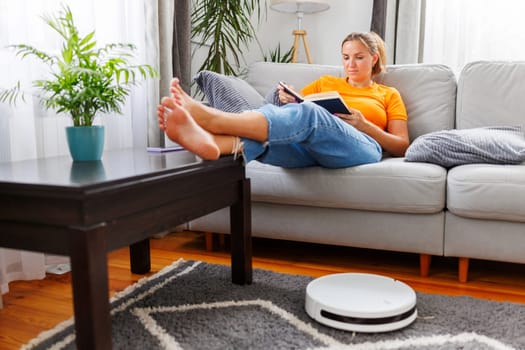 A woman enjoys some downtime reading a book on the couch with her feet up while a robot vacuum cleaner works in the background.