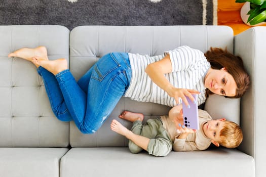 An aerial view of a relaxed mother and her young son lying on a couch, taking a selfie together