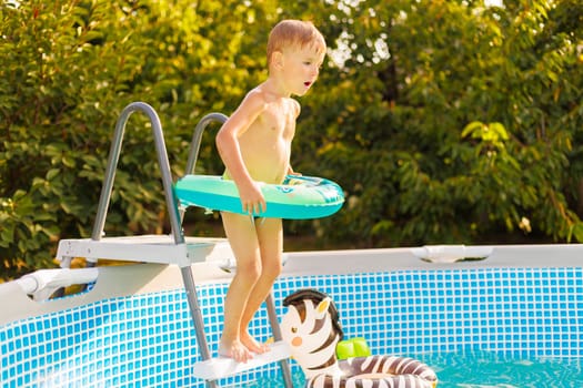 A young boy holding a float ring stands by a swimming pool ladder, excited and ready to jump in for a swim on a sunny day.