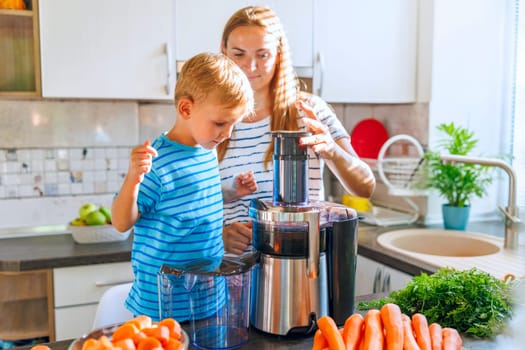 A young boy and his mother prepare homemade juice with fresh carrots using an electric juicer in a sunny kitchen.