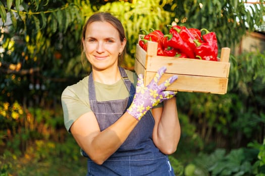 A happy woman in a garden smiles while holding a wooden crate filled with fresh red peppers, showcasing homegrown produce.
