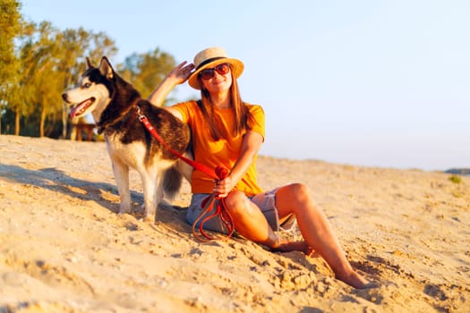 A woman in a straw hat and sunglasses relaxes on the beach with her Siberian Husky, enjoying a beautiful sunset together.