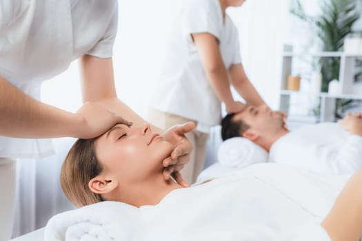 Caucasian couple enjoying relaxing anti-stress head massage and pampering facial beauty skin recreation leisure in dayspa modern light ambient at luxury resort or hotel spa salon. Quiescent