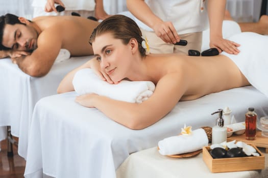 Hot stone massage at spa salon in luxury resort with day light serenity ambient, blissful couple customer enjoying spa basalt stone massage glide over body with soothing warmth. Quiescent