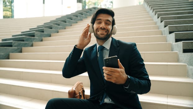 Smiling business man listening music while looking at mobile phone. Project manager holding phone while enjoy hip-hop song at stairs. Caucasian leader listen relax sound while plan project. Exultant.