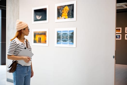 Young person at photo frame hold digital book leaning against at show exhibit artwork gallery, Asian woman holding tablet at art gallery collection in front framed paintings looking pictures on wall