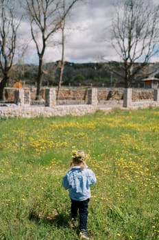 Little girl in a wreath stands on a flowering lawn and looks at a stone fence. Back view. High quality photo