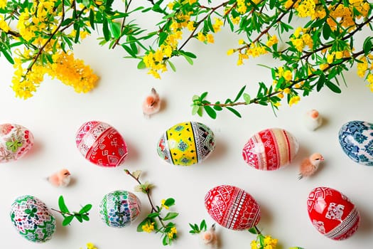 An aerial view of the Easter eggs lying on a white ground. Next to the Easter eggs are little chickens and fresh twigs from the spring trees.