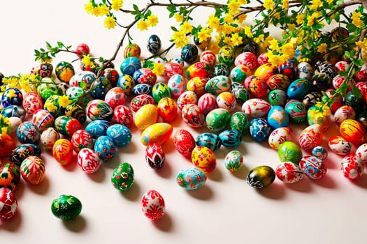 Easter eggs in various shades and colours lie on a white tabletop. An aerial view of a large number of colourful Easter eggs. A branch plucked from a forsythia bush can be seen next to the eggs.