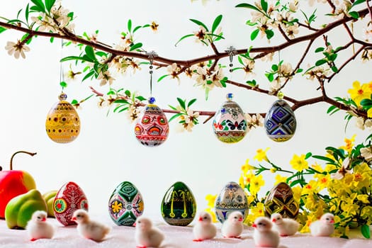 A branch plucked from a flowering spring tree. There are white flowers on the branch and a painted Easter egg hanging from it. At the bottom stands a row of Easter eggs, yellow flowers and white chickens.
