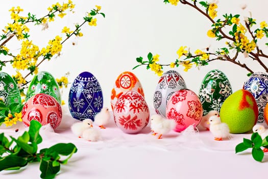 Little white chickens walk among the colourful Easter eggs. The eggs are set against a white background. At the back you can see some branches with small yellow flowers.