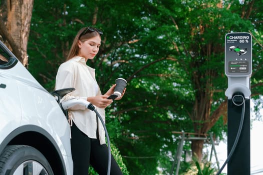 Young woman with coffee cup and sustainable urban commute with EV electric car recharging at outdoor cafe in springtime garden, green city sustainability and environmental friendly EV car. Expedient