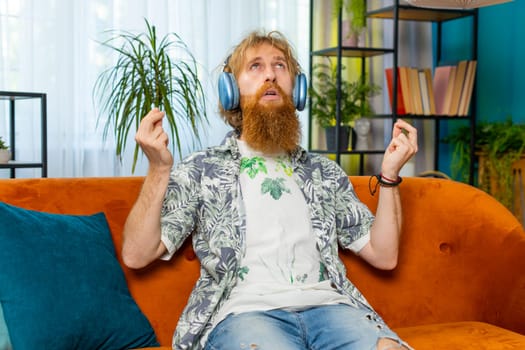 Keep calm down, relax. Caucasian redhead man listening music breathes deeply, eyes closed meditating with concentrated thoughts, peaceful mind. Young bearded guy sits at home in living room on couch