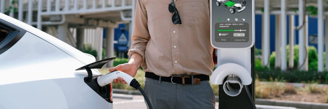 Young man recharge electric car's battery from charging station in city commercial parking lot. Rechargeable EV car for sustainable environmental friendly urban travel. Panorama Expedient