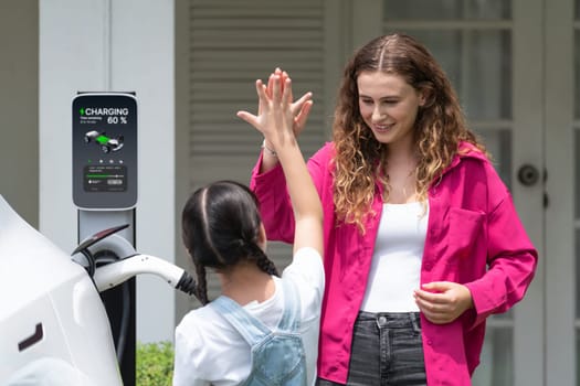 Happy little young girl learn about eco-friendly and energy sustainability as she help her mother recharge electric vehicle from home EV charging station. EV car and modern family concept. Synchronos