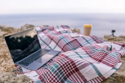 Laptop coffee on blanket with ocean view. Illustrating serene outdoor laptop use. Freelancer enjoying their time outdoors while working or browsing the internet