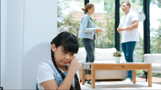 Stressed and unhappy young girl huddle in corner, cover her ears blocking sound of her parent arguing in background. Domestic violence at home and traumatic childhood develop to depression. Synchronos