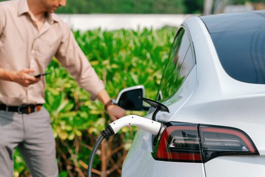 Young man use smartphone to pay for electricity at public EV car charging station green city park. Modern environmental and sustainable urban lifestyle with EV vehicle. Expedient